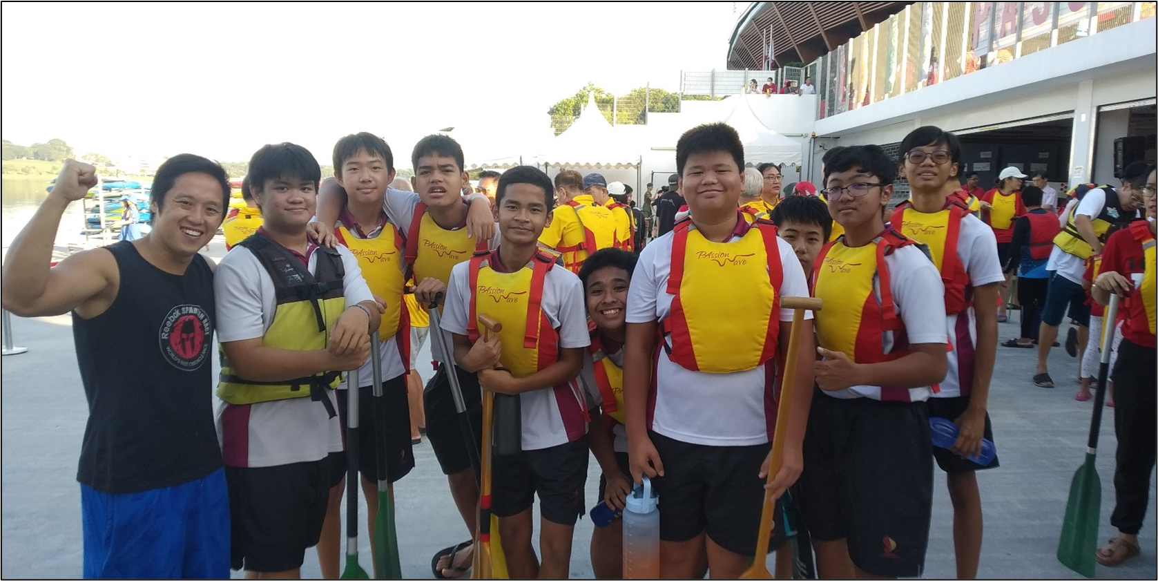 A group of young male dragon boat paddlers in yellow life vests pose together holding paddles near a waterfront venue.